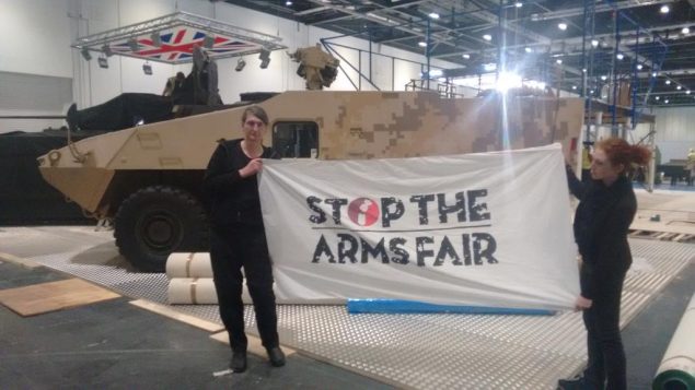 Two activists hold a 'Stop the Arms Fair' banner in front of an armoured vehicle