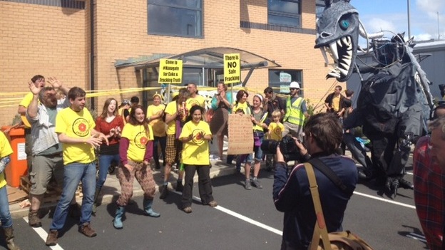 Crowd of protesters dancing outside a beige brick building with banners. One reads 'There is NO safe fracking'. There is a giant grey monster/dinosaur prop. 