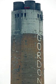 A power station chimney with 'Gordon' written in white down the side. 5 Greenpeace protesers are at the top of the chimney in red jumpsuits and hard hats. 