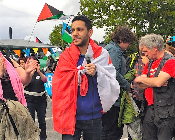 A man draped in a Bahraini flag holds a microphone as a Palestine flag waves in the background.