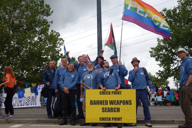 Veterans in blue hoodies and t-shirts, holding Peace and Palestine flags, hold a big yellow sign that reads, 'Check point - Banned weapons search - Check point'