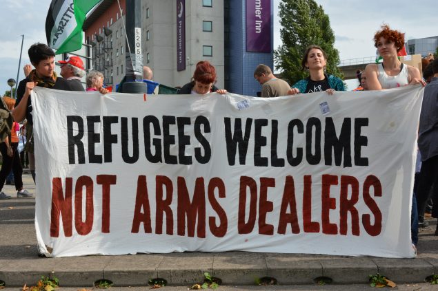 4 people hold a banner that reads, Refugees Welcome Not Arms Dealers