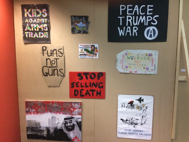 Protest art on a wall, hand-painted signs saying Puns not guns, Peace Trumps War, Stop Selling Death; stencil for Kids Against Arms Trade, Pastel-coloured butterfly with hand-drawn letters saying Welfare not Warfare; Collage featuring Saudi Prince bin Salman, target symbols and words ‘the war in Yemen’ and PROTEST; Dove collage; Printed placard saying Stop Arming Human Rights Abusers