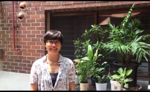 Jungmin, wearing a white patterned shirt, stands in front of a black, metal garage door set in a brick wall behind several plants growing in pots