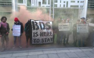 A group of people stand with banners saying 'BDS is here to stay' in front of red smoke
