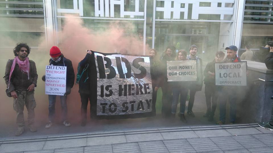 A group of people stand with banners saying 'BDS is here to stay' in front of red smoke