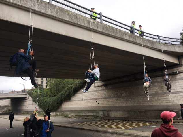 Four abseilers are suspended from a road bridge, blocking the road. Police officers stand above them. 