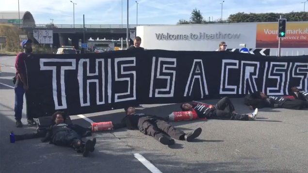 5 people lie in the road in arm tubes, three more people stand behind them with a banner that reads, 'This is a Crisis'
