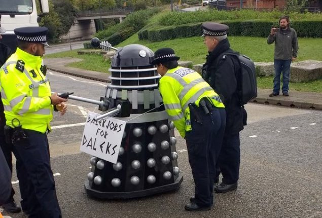 Police officers interrogate a dalek in the road. The dalek has a banner that reads, "Dalek Jobs for Daleks"