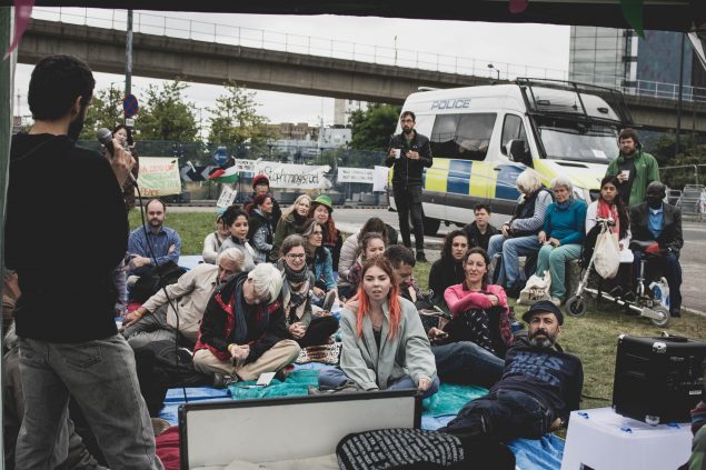 A workshop runs on the grass beside the roundabout. People sit listening to a speaker, there is a police van behind them and banners in the background. 