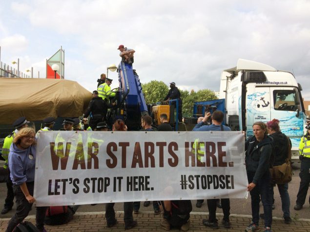 Two people hold a banner that says, War Starts Here Let's Stop It Here #StopDSEI in front of a truck carrying equpiment which is stopped in the road. Someone is sat on top of the equipment. 