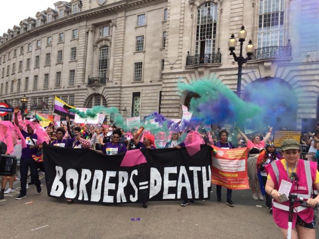 Black people and people of colour hold up a banner saying "Borders = Death" while setting off blue smoke