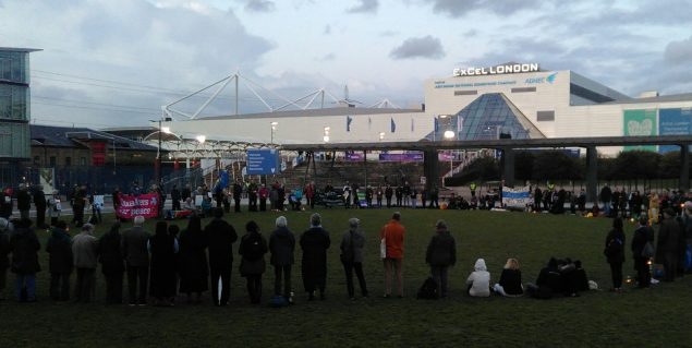 People standing in a circle at dusk outside the ExCel Centre with candles and banners