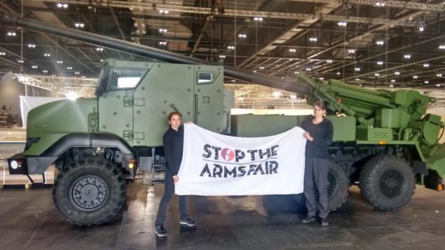 Two activists hold a 'Stop the Arms Fair' banner in front of a dark green armoured vehicle